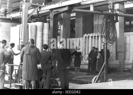 Slawjansk, 1983. Gebiet des Armature-Isolator Plant (AIZ). Schwarzweiß-Archivfoto, das eine faszinierende historische Szene einfängt. Mehrere fleißige Fabrikarbeiter, darunter Männer in Arbeitskleidung und eine Frau in dunklem Kleid, versammeln sich in einer geräumigen Werkstatt. Sie stehen neben einem schweren Ofenwagen, beladen mit weißen Keramikzylindern, unter einem komplexen Deckenkran, der für die Fertigung verwendet wird. Dieses detaillierte Bild bewahrt ein echtes Fragment der sowjetischen Industriegeschichte und zeigt das Leben vor dem Krieg und den friedlichen Donbass. Stockfoto