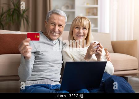 Seniorenpaar zeigt Kreditkarte, während er zu Hause mit Laptop sitzt Stockfoto