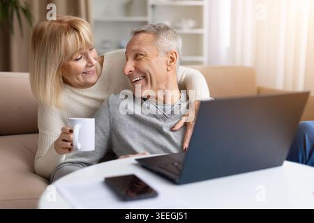 Glückliches Seniorenpaar, das einen Laptop benutzt, während er sich zu Hause bei einem Kaffee entspannt Stockfoto