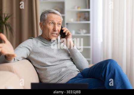 Schockierter reifer Mann, der zu Hause auf dem Sofa sitzt und mit seinem Smartphone spricht Stockfoto