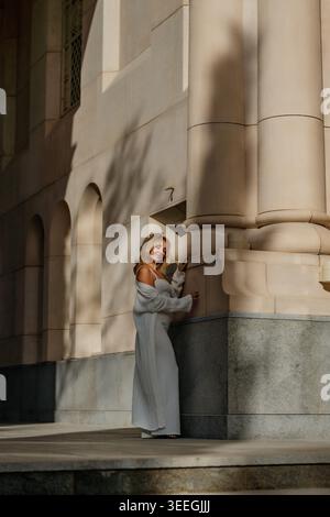 Frau, Gebäude, Schatten - Frau in einem langen grauen Kleid steht an einer Steinsäule, die einen Schatten auf eine Wand wirft. Stockfoto