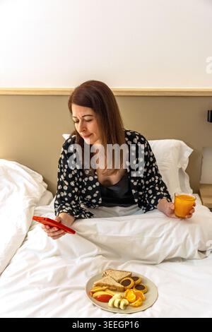Frau genießt Frühstück im Bett, während sie telefoniert Stockfoto