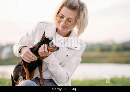 Glückliche Frau, die ihren russischen Toy Terrier draußen kuschelt. Kleiner schwarzer und brauner Hund in den Händen des Besitzers, Tierpflege und Bindungskonzept in einem Park, Lifestyle-Porträt der Miniatur-Rasse und ihres Besitzers, Freundschaft Stockfoto