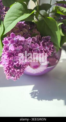 Ein Blumenstrauß aus blühenden Fliedern mit grünen Blättern in einer runden Glasvase mit Blumenmuster. Ein Frühlings-Blumenarrangement in einem Innenraum mit weicher Natur Stockfoto