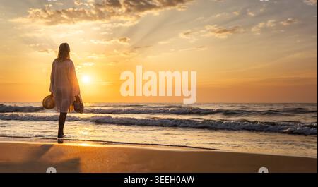 Junge Frau, die bei Sonnenuntergang am Sandstrand steht und Sandalen und Hut hält, während sie die Wellen des Meeres in der Abenddämmerung beobachtet Stockfoto