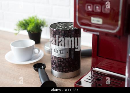 Nahaufnahme eines dekorativen Kaffeebohnenbehälters neben einer roten Espressomaschine und einer weißen Kaffeetasse auf einer Küchenarbeitsfläche mit natürlichem Licht und Schiefer Stockfoto