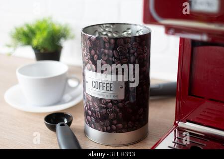 Nahaufnahme eines dekorativen Kaffeebohnenbehälters neben einer roten Espressomaschine und einer weißen Kaffeetasse auf einer Küchenarbeitsfläche mit natürlichem Licht und Schiefer Stockfoto