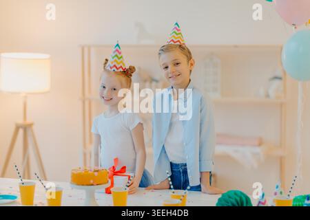 Zwei Kinder tragen Partyhüte, die zu Hause neben der Geburtstagskuche stehen und einen Geschenktisch tragen Stockfoto