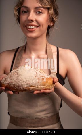 Selbstbewusste junge Bäckerin, die frischen handwerklichen Brotlaib hält und stolz hausgemachte Backwaren präsentiert Stockfoto