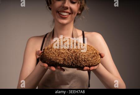 Junge Bäckerin, die ein frisch gebackenes dunkles Brot mit Sesamsamen hält und präsentiert, lächelnd Stockfoto