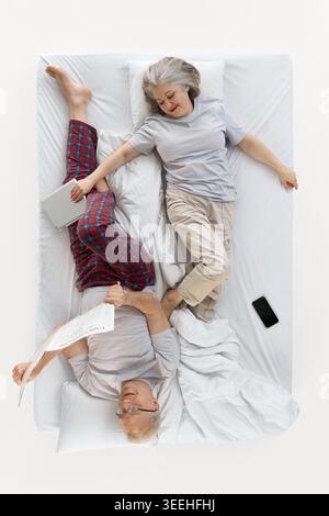 Eine ältere Frau, die neben einem Mann lag, der Zeitung las, in einem weißen Bett von oben. Stockfoto