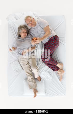 Seniorenpaar teilen sich den Smartphone-Bildschirm, während sie zusammen im weißen Bett liegen. Stockfoto