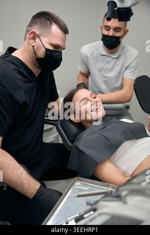 Junge Frau lächelt im Zahnarztstuhl mit Fachleuten während der Prüfung Stockfoto