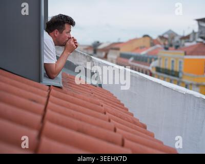 Gestresster oder müder Mann, der sich aus einem Dachfenster beugt und den Kopf in den Händen hält Stockfoto