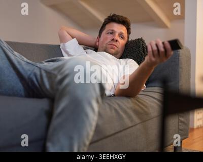 Erschöpfter Mann, der auf einem grauen Sofa schläft, während er eine Fernbedienung hält Stockfoto