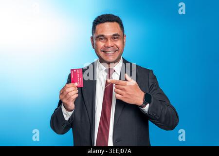 Lächelnder indonesischer Mann in schwarzem Anzug und Krawatte, der eine Kreditkarte hält und darauf zeigt. Studio-Aufnahme auf blauem Hintergrund für Finanz- und Bankwesen. Stockfoto