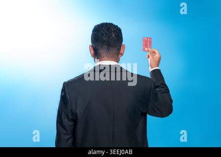 Rückansicht eines indonesischen Mannes in einem Anzug, der eine rote Kreditkarte vor blauem Hintergrund hält. Konzept des finanziellen Erfolgs und des Premium-Zugangs. Stockfoto