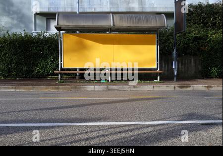 Bushaltestelle mit leerer Plakatwand und Stoppstange am Straßenrand. Hecke und Appartementgebäude hinten, Straße davor. Hintergrund für Kopierbereich. Stockfoto