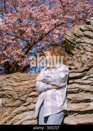 Die junge Frau lehnt sich im Frühjahr an alten Baumstamm unter rosa Kirschblüten in Tallinn, Estland Stockfoto