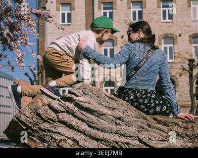 Zwei Kinder klettern auf einem großen Holzstamm in einem Stadtpark von Tallinn mit Kirschblüten und Steinbau im Frühjahr Stockfoto