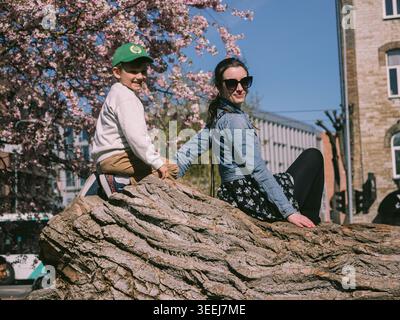 Frau und Junge sitzen auf einem großen Baumstamm unter blühenden Kirschblüten in Tallinn, Estland, im Frühjahr Stockfoto