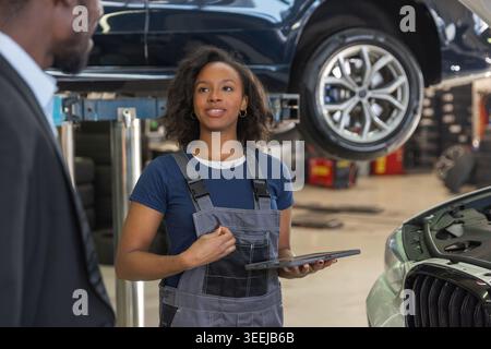 Professionelle Mechanikerin in einer modernen Werkstatt, die ein Tablet benutzt, um einem Kunden, einem afroamerikanischen Geschäftsmann, eine Panne zu erklären. Stockfoto