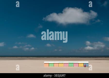 Pastellfarbene Strandhütten am MEER, die allein am riesigen Sandstrand unter blauem Himmel mit verstreuten weißen Wolken stehen, Paare und Sonnenschirme entspannen. Stockfoto