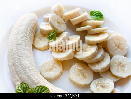 Blick von oben auf eine ganze geschälte Banane und frische Scheiben auf weißem Hintergrund, dekoriert mit grünen Minzblättern. Stockfoto