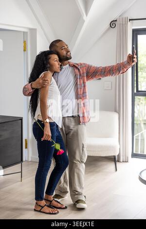 Diverse Paare, die Selfie im hellen Wohnbereich machen, Mann mit Smartphone, Frau mit roter Rose Stockfoto