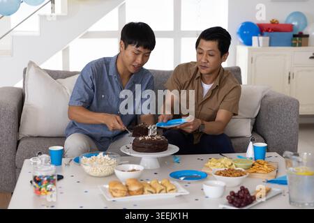 Zwei asiatische Männer sitzen auf einem grauen Sofa im Wohnzimmer und schneiden Schokoladenkuchen mit numerischer Kerze Stockfoto