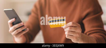 Person, die ein Smartphone nutzt und eine gelbe Kreditkarte in einem zwanglosen Ambiente in Innenräumen hält Stockfoto