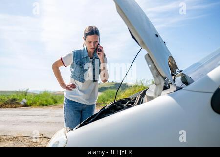 Frau neben einem Pannenwagen mit offener Motorhaube, die an einem sonnigen Tag während der Reise einen Anruf für Pannenhilfe tätigt Stockfoto