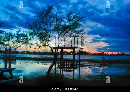Intensiver blauer und orangener Abendhimmel über einer ruhigen Lagune. Die Silhouette eines Baumes und ein hölzerner Pavillon heben sich von den lebhaften Farben der Wolken ab Stockfoto