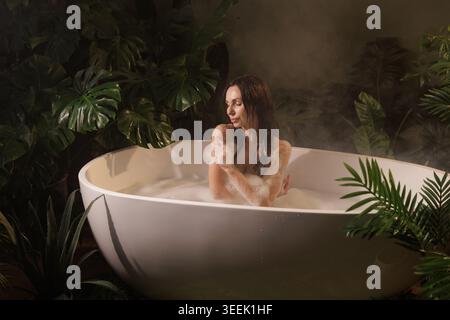 Eine ruhige Szene einer Frau, die ein Sprudelbad in einer modernen Badewanne genießt, umgeben von üppigem tropischem Grün, perfekt für Wellness- und Schönheitskampagnen. Stockfoto