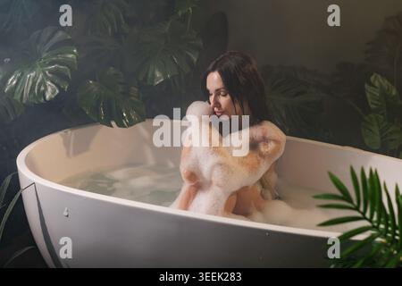 Eine ruhige Frau, die ein Sprudelbad in einer modernen Badewanne genießt, umgeben von üppigem tropischem Laub, perfekt für Beauty- und Wellness-Kampagnen. Stockfoto