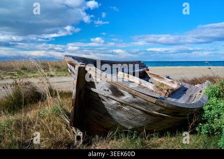 Ein altes verlassenes hölzernes Fischerboot, das an einem grasbewachsenen Ufer nahe einem Sandstrand am Meer unter einem blauen bewölkten Himmel verfällt. Stockfoto