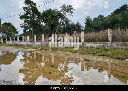 Eine große schmutzige Wasserpfütze reflektiert auf einer unbefestigten Straße vor einem alten Betonzaun mit Säulen und grünen Kiefern. Stockfoto