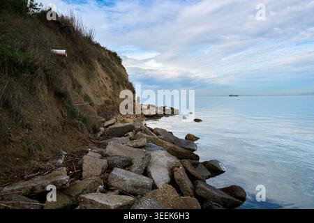 Küstenerosion mit einer steilen Lehmklippe und Steinblöcken an einem felsigen Strand vor einer ruhigen Meereswasseroberfläche unter bewölktem Himmel. Stockfoto