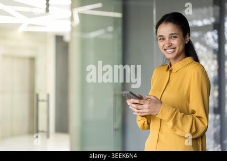 Eine junge Geschäftsfrau, die in einem modernen Bürokorridor steht, auf die Kamera blickt und lächelt, während sie ihr Smartphone hält, was für Kommunikation und Konnektivität steht Stockfoto