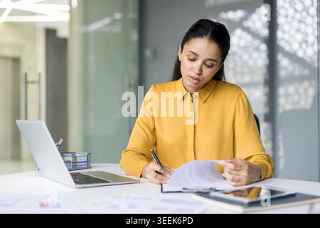 Junge professionelle Frau, die Dokumente überprüft und Notizen an ihrem Schreibtisch macht, zeigt Konzentration und Hingabe für Aufgaben in einer modernen Büroumgebung Stockfoto