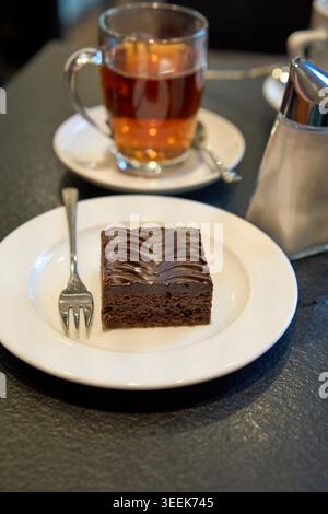 Ein quadratisches Stück Schokoladenkuchen auf einem Teller mit einer Gabel, serviert mit einer heißen Tasse schwarzen Tee und einem Zuckerstreuer in einem Café. Stockfoto