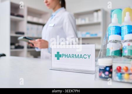 Junge Frau Apotheke mit Tablette hinter Apotheke Theke mit Medizin Display, repräsentiert Einzelhandel Gesundheitswesen Service. Ideal für Rezepte, Medikamente, klinisches Marketing Stockfoto