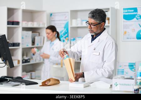 Indischer Apotheker mittleren Alters, der verschreibungspflichtige Medikamente an der Apotheke verpackt, während eine Kollegin im Hintergrund die Bestellung vorbereitet. Ideal für den Einzelhandel im Gesundheitswesen und das Marketing von Medikamentendienstleistungen Stockfoto