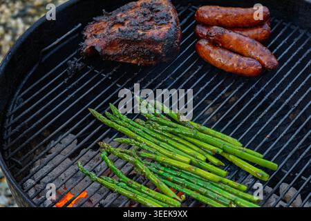 Verschiedene gewürzte Fleischsorten und frische grüne Spargelspeere werden über glühender heißer Holzkohle gegrillt Stockfoto