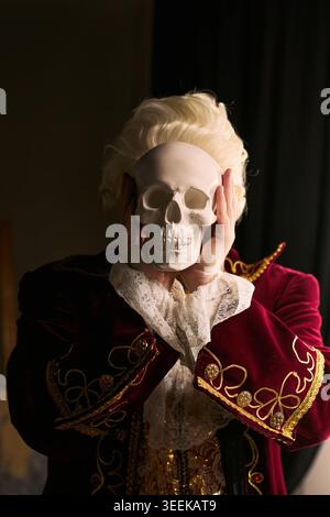 Vertikale Aufnahme des Darstellers, der den Schädel über das Gesicht hält, während er in historischem Kostüm posiert, was die Sterblichkeit und das Theaterdrama symbolisiert. Ideal für Bühnenwerbung, Hamlet-Konzept, Event-Marketing Stockfoto