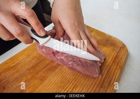 Nahaufnahme von weiblichen Händen, die frisches rohes Schweinefilet mit weißem Keramikmesser auf hölzernem Schneidebrett in der Küche schneiden Stockfoto