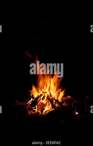 Große Lagerfeuer-Flammen, die aus gestapeltem Holz in völliger Dunkelheit aufsteigen. Das Bild erfasst vertikale Bewegungen, intensive orangene Hitze und Funken in der Luft Stockfoto