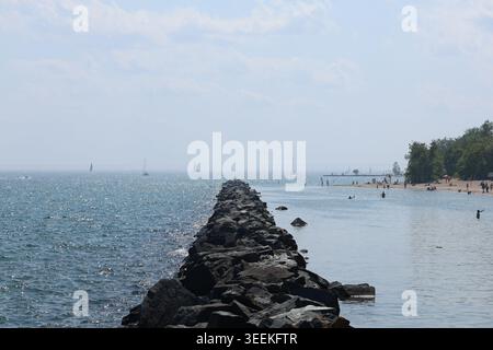 Steinbruch, der in den Lake Ontario führt, mit Segelbooten und Strandgängern, die in der Ferne sichtbar sind. Fotografiert an einem hellen Sommertag mit blauem Wasser und Stockfoto