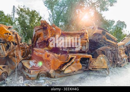 Ein großer Haufen vollständig verbrannter und rostfreier Metallwagen, die durch ein Feuer zerstört wurden, stand draußen in der Nähe von grünen Bäumen mit starken Sonnenstrahlen Stockfoto
