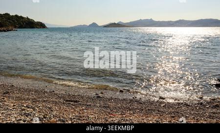 Kieselstrand mit Blick auf die glitzernde Ägäis in Bitez, Bodrum. Sonnendurchflutetes Wasser sorgt für eine entspannte Sommerferienszene Stockfoto